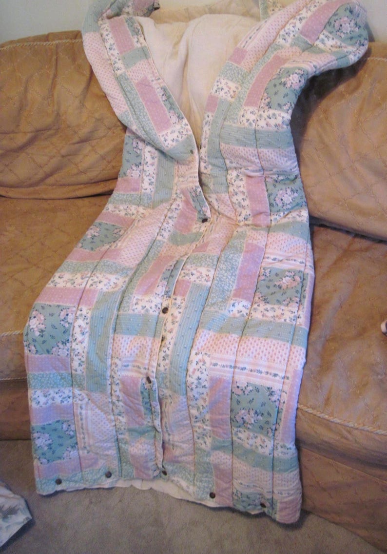 May include: A quilted robe in pastel colors of pink, green, and white, with a floral pattern. The robe has a button-up front and a shawl collar. The robe is lying on a brown couch.
