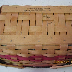 Pu&ograve; includere: Un cesto rettangolare Longaberger con un design intrecciato. Il cesto &egrave; realizzato in legno marrone chiaro con una fascia rosa. La parte superiore del cesto riporta la scritta "LONGABERGER BASKETS HANDWOVEN DRESDEN, OHIO USA".