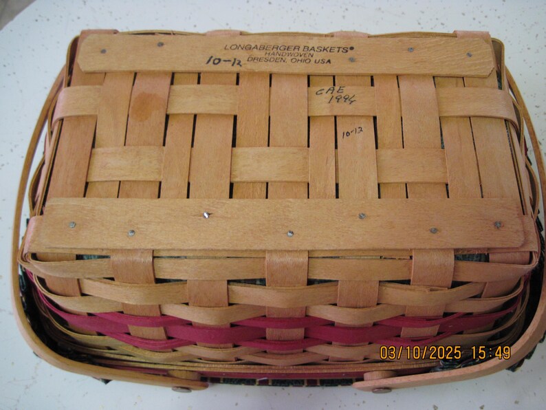 Pu&ograve; includere: Un cesto rettangolare Longaberger con un design intrecciato. Il cesto &egrave; realizzato in legno marrone chiaro con una fascia rosa. La parte superiore del cesto riporta la scritta "LONGABERGER BASKETS HANDWOVEN DRESDEN, OHIO USA".