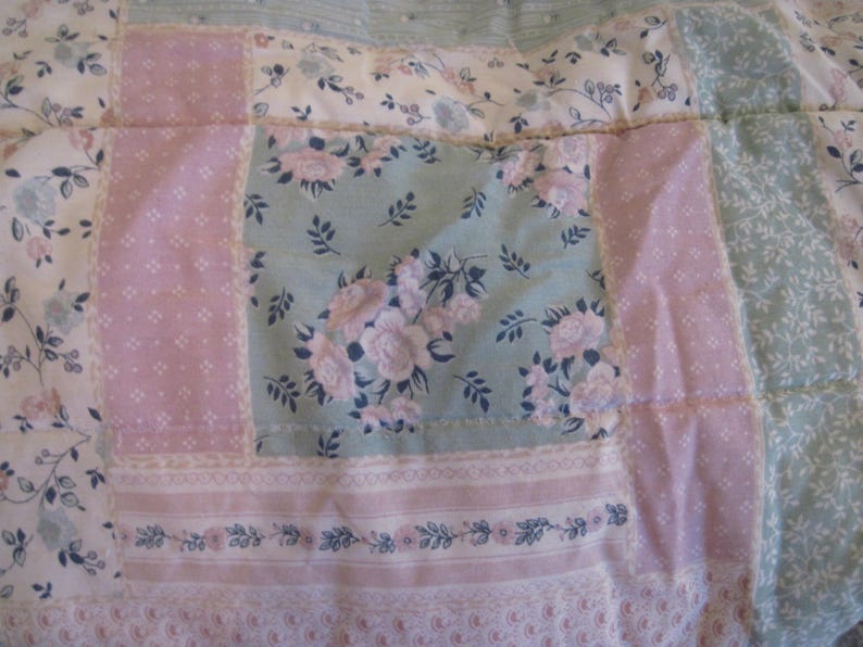May include: Close-up of a patchwork quilt with floral and geometric patterns. The quilt features squares of light pink, pale green, and white fabric. Some squares have floral prints, while others have small dot patterns. The quilt is a mix of soft colors and textures.