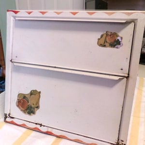 May include: Vintage white metal bread box with two compartments. The box has a red and orange trim and fruit decals. The box shows signs of wear and tear, adding to its vintage charm.