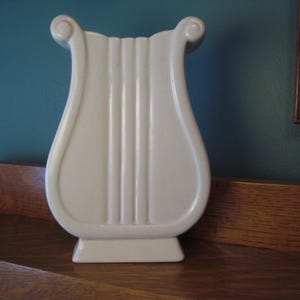 May include: A white ceramic vase in the shape of a lyre, with vertical grooves down the front and scrolled accents at the top. The vase is displayed on a wooden surface against a teal wall.