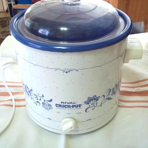 May include: White Rival Crock-Pot with a blue lid and trim. The slow cooker has blue floral designs and the words "RIVAL CROCK-POT" printed on the front. It has a white power cord and a dial with settings for Off, Low, and High.
