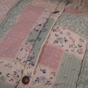 May include: Close-up of a quilted jacket with a floral pattern. The jacket features a patchwork design with pink, white, and light green floral and dotted fabric panels. A decorative button secures the jacket.