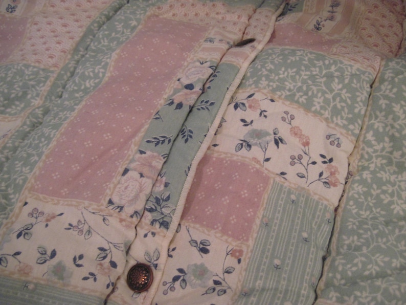 May include: Close-up of a quilted jacket with a floral pattern. The jacket features a patchwork design with pink, white, and light green floral and dotted fabric panels. A decorative button secures the jacket.