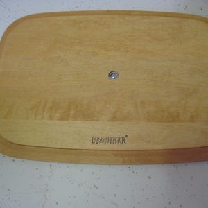 Pu&ograve; includere: Un vassoio da portata in legno marrone chiaro con angoli arrotondati. Il vassoio ha un'area incassata e un elemento di fissaggio in metallo al centro. Il logo Longaberger Woodcrafts &egrave; visibile in basso.
