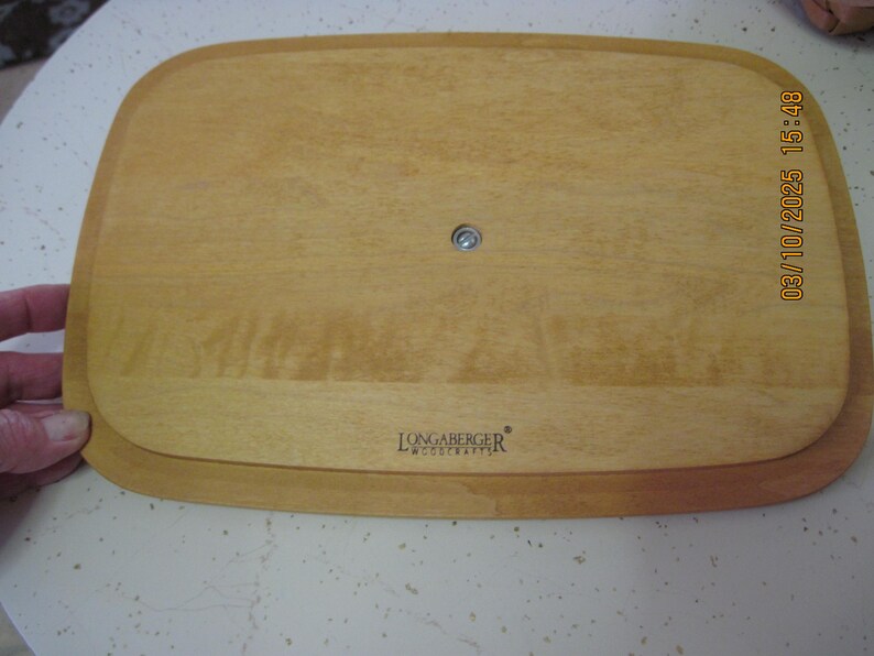 Pu&ograve; includere: Un vassoio da portata in legno marrone chiaro con angoli arrotondati. Il vassoio ha un'area incassata e un elemento di fissaggio in metallo al centro. Il logo Longaberger Woodcrafts &egrave; visibile in basso.