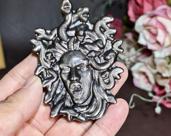May include: A detailed, antique-style Medusa head sculpture in a metallic silver finish. The sculpture features intricate details of Medusa's face and the snakes that form her hair. The piece is held in a hand, showcasing its size and craftsmanship.