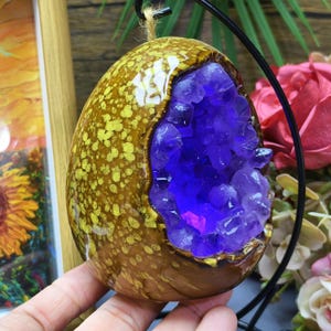 May include: A decorative, egg-shaped ornament with a brown and yellow speckled exterior. The center reveals a vibrant purple crystal geode. The ornament is suspended by a black wire and twine.