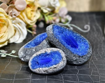 May include: Three oval-shaped stones with a rough, gray exterior and a vibrant blue, crystal-like interior. The stones are arranged on a textured, dark surface, with a blurred floral arrangement in the background.
