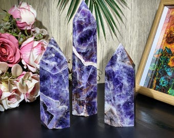 May include: Three amethyst crystal towers in varying sizes. The crystals display a deep purple hue with white and brown veining. The crystals are displayed in front of a floral arrangement and a framed painting.