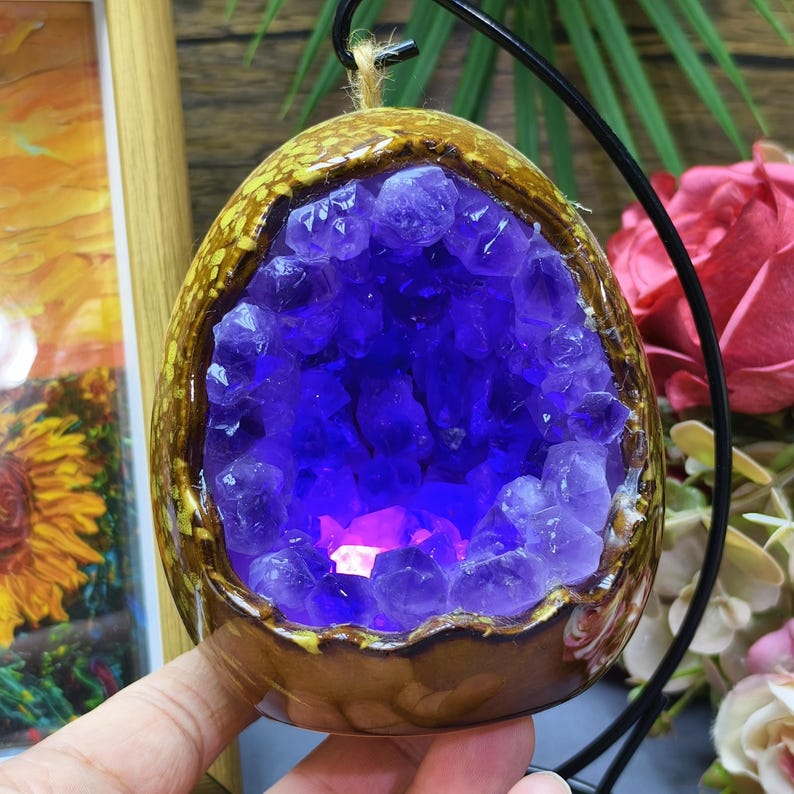 May include: A ceramic geode-shaped ornament with a deep purple amethyst crystal interior. The exterior is brown with a yellow-gold trim. A black metal stand holds the ornament, and a natural twine loop is at the top.