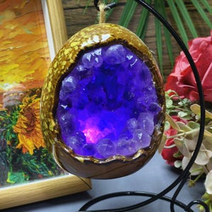 May include: A decorative, egg-shaped ceramic geode with a vibrant purple amethyst crystal interior. The exterior is a mottled brown and yellow, with a dark brown rim. The geode hangs from a black metal stand, with a painting in the background.