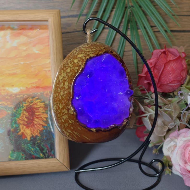May include: A decorative, egg-shaped lamp with a brown, textured exterior and a vibrant blue interior. The lamp is suspended from a black metal stand. A painting in a wooden frame is visible in the background.