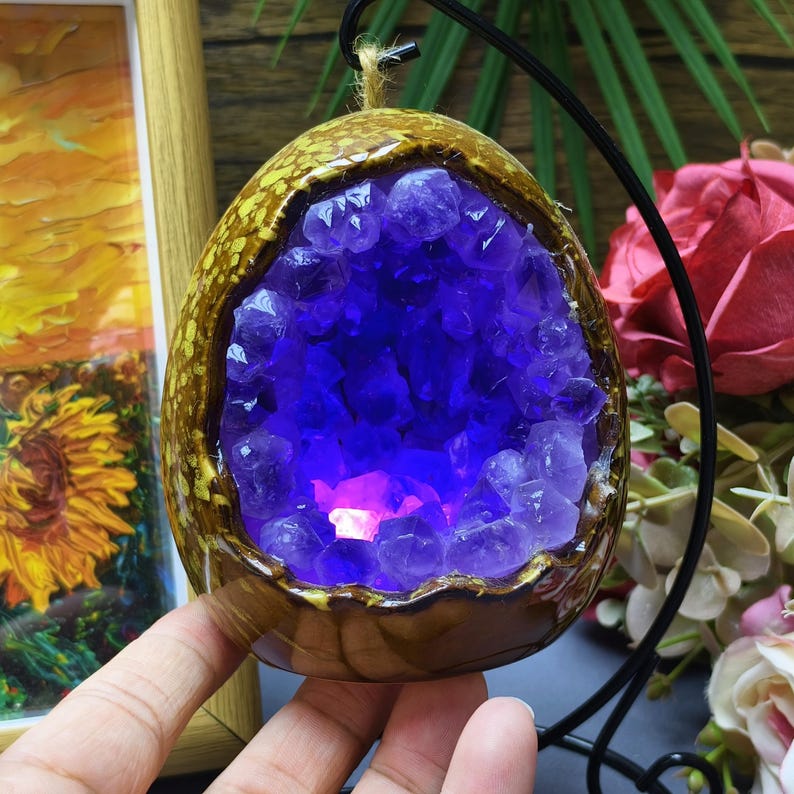 May include: A decorative, egg-shaped amethyst geode pendant. The geode is encased in a brown, textured ceramic shell with a golden rim. The interior is filled with vibrant purple crystals, illuminated by a soft, pinkish light.