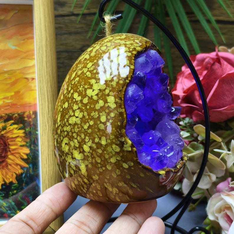 May include: A decorative, egg-shaped geode with a brown and yellow speckled exterior. The geode has a large opening revealing a cluster of vibrant purple amethyst crystals. The geode is suspended from a black metal stand.