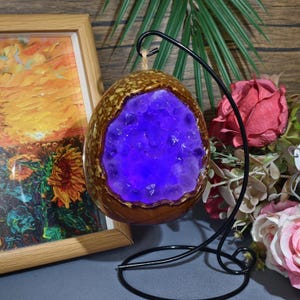 May include: A decorative, egg-shaped object with a brown, textured exterior and a vibrant purple, crystal-filled interior. The object is suspended from a black metal stand. A framed painting with sunflowers and a sunset is in the background.