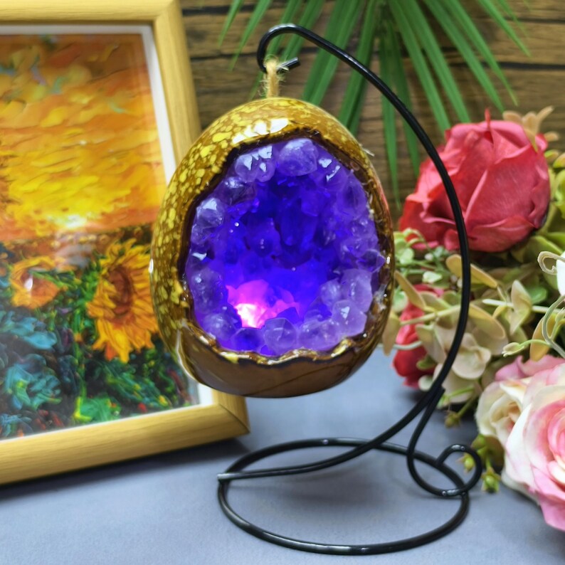 May include: A decorative, egg-shaped amethyst geode with a golden-brown exterior, suspended from a black metal stand. The geode's interior is filled with vibrant purple crystals, illuminated by a soft, pinkish light.