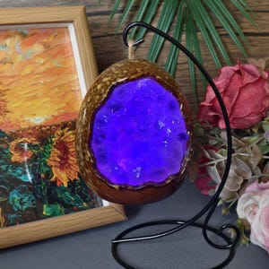 May include: A brown, egg-shaped decorative object with a glowing purple interior, displayed on a black metal stand. The object has a textured, speckled surface and a rough, open center. A framed painting of sunflowers is in the background.