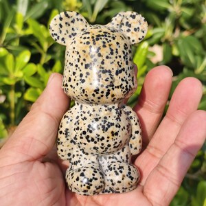 Könnte beinhalten: Gefleckte Bärenfigur mit cremefarbenem Grund und schwarzen Punkten. Der Bär hat abgerundete Ohren, einen kleinen Körper und stämmige Beine. Die Figur wird in einer Hand vor grünem Laub gehalten.