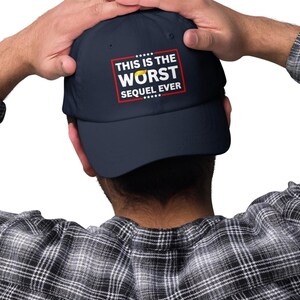 Pu&ograve; includere: Cappellino da baseball blu navy con la scritta "THIS IS THE WORST SEQUEL EVER" in bianco e rosso. Il cappellino ha una visiera curva e una corona strutturata. Il cappello &egrave; indossato.