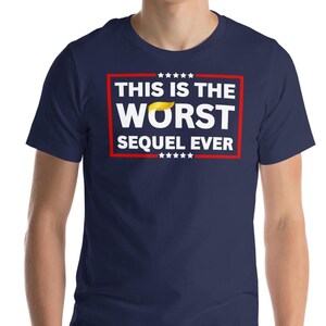 May include: Navy blue t-shirt with the text "THIS IS THE WORST SEQUEL EVER" in a white and red rectangular design. The design includes a yellow hair graphic. The shirt is a crew neck style.
