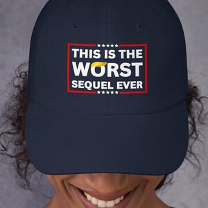 Pu&ograve; includere: Cappellino da baseball blu navy con la scritta "THIS IS THE WORST SEQUEL EVER" in lettere bianche all'interno di un rettangolo rosso. Il cappellino presenta un piccolo disegno di capelli gialli. Il cappello &egrave; indossato da una persona.