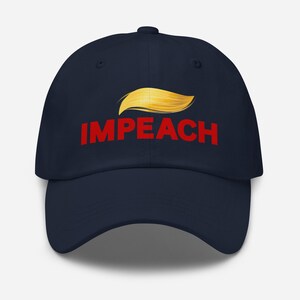 May include: A navy blue baseball cap with the word "IMPEACH" in red block letters. Above the word is a stylized golden-yellow hair design. The cap has a curved brim and a fabric strap closure.