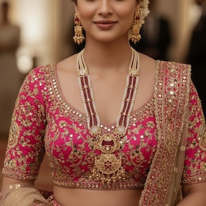 Puede incluir: Una mujer con una blusa rosa bordada y una falda a juego, adornada con joyas de oro. El collar presenta un colgante grande y se complementa con pendientes a juego. El atuendo es adecuado para una boda u ocasión especial.