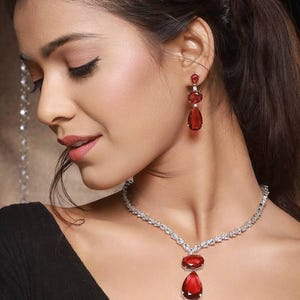 May include: A diamond necklace and matching earrings with large, teardrop-shaped red gemstones. The necklace and earrings are set in a silver-coloured metal. The jewellery is worn with a black top.