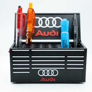 May include: Black desk organizer with the Audi logo and a toolbox design. The organizer holds pens and a marker. The Audi logo is white and red. The organizer has a series of horizontal white lines.