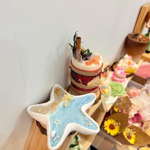 May include: A white starfish-shaped ornament with a blue and sand-coloured interior, decorated with pearls and small star accents. A cylindrical candle with floral and cinnamon stick accents is behind it.