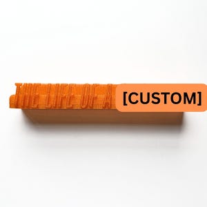 May include: An orange rectangular stamp with the text "HELLELUJA" carved into the top. A rectangular orange sticker with the word "[CUSTOM]" is attached to the side. The stamp is set against a white background.