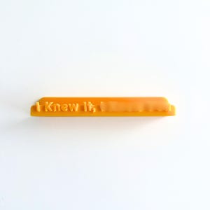 May include: A rectangular orange plastic stamp featuring the embossed text "I Knew It," followed by a blurred word. The object has a flat base and a raised handle section, designed for creating impressions with a consistent, bold, and sans-serif font.