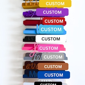May include: A vertical arrangement of colorful, custom-made labels. Each label has a different color, including green, yellow, purple, red, blue, black, pink, gray, brown, and orange. The word "CUSTOM" is printed in white on each label. Some labels have additional text.