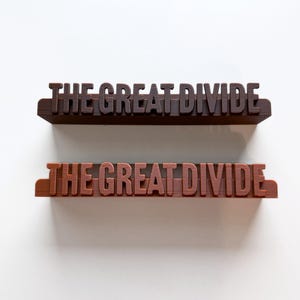 Puede incluir: Dos estantes impresos en 3D con el texto "THE GREAT DIVIDE". Uno es marrón oscuro, el otro marrón rojizo. Ambos están sobre un fondo blanco. Adecuado para la decoración.