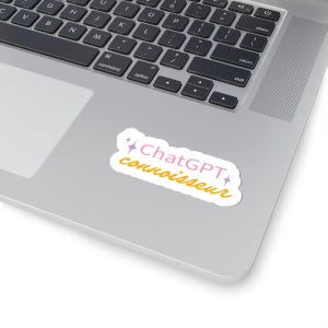 May include: A white sticker with pink and yellow text that reads "ChatGPT connoisseur" with two pink stars on either side of the text. The sticker is on a silver laptop.