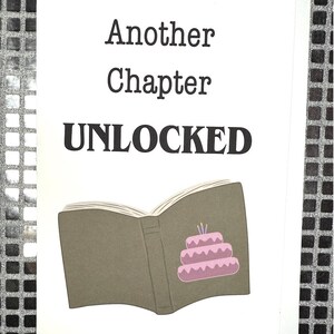 Pode incluir: Um cartão branco com texto preto que diz "Another Chapter Unlocked". Um livro castanho com um bolo rosa com velas dentro está ilustrado no cartão.