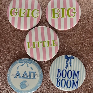 May include: Five round buttons with various designs. Three buttons have pink and white stripes with the words "GBIG", "BIG", and "LITTLE" in green. Two buttons are blue and white, one with Greek letters and a lion, the other with "BOOM BOOM".