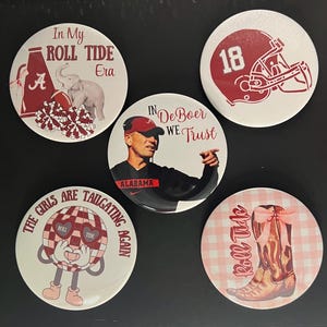 May include: Five round buttons with different designs related to the University of Alabama Crimson Tide football team. The buttons feature a football helmet, a coach, a cheerleader, a football, and a boot.
