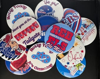 May include: A collection of round pin-back buttons featuring various designs in red, white, and blue. The buttons display text including Hotty Toddy, Meet you at the Oxford Tailgate, REBELS, Ole Miss, and Mississippi, alongside graphics of cowboy hats, footballs, and mascots.