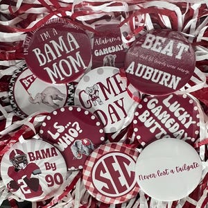 May include: A collection of burgundy and white game day buttons with various phrases and graphics. The buttons include text like "Bama Mom," "Beat Auburn," and "Lucky Gameday Butt." The buttons are surrounded by red and white paper filler.
