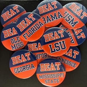 May include: A collection of round pin-back buttons featuring a split blue and orange design. Each button displays the word BEAT in orange or white text above the name of a university team, including Alabama, Florida, Ole Miss, LSU, Georgia, and others.