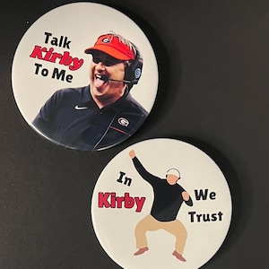 May include: Two white round buttons. One button features a photo of a man wearing a red visor and the text "Talk Kirby To Me". The other button has an illustration of a man and the text "In Kirby We Trust".