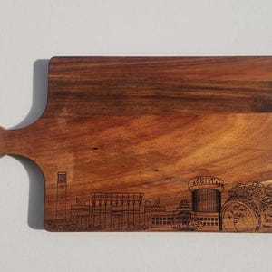 Puede incluir: Una tabla de cortar rectangular de madera con asa. La tabla está grabada con un horizonte de edificios y el texto "WELCOME TO AGGIELAND" y "Texas A&M Home of the 12th Man".