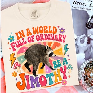 May include: A cream-colored t-shirt featuring a graphic of a raccoon centered within a colorful sunburst design. The text reads, "IN A WORLD FULL OF ORDINARY BE A JIMOTHY," surrounded by illustrations of mushrooms, flowers, and lightning bolts.