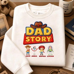 May include: White sweatshirt with a colourful embroidered "DAD STORY" design, featuring cartoon characters and names. The image includes a coffee mug, a teddy bear, and a wooden block that says "BEST DAD".