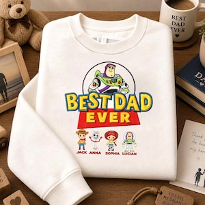 May include: White sweatshirt with the text "BEST DAD EVER" and Toy Story characters. The sweatshirt has a crew neck and long sleeves. A mug with "BEST DAD EVER" is also visible, along with a teddy bear and a wooden block.