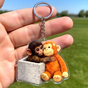 May include: A clear acrylic keychain featuring two cartoon monkeys. One brown monkey is in a gray box, hugging an orange monkey. The keychain has a silver ring and chain. The background is a green field and blue sky.