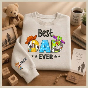May include: A light grey sweatshirt with the text "Best Dad Ever" in a colourful design. The sleeve features the name "Jack" and a cartoon cowboy. A teddy bear, framed photo, and coffee mug are also visible.